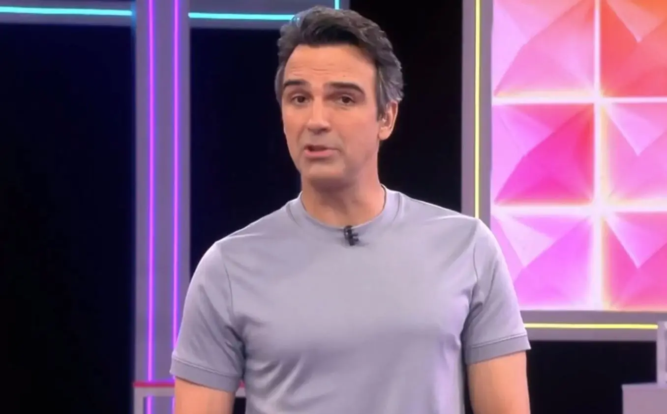 Pessoa em pé em um estúdio iluminado, vestindo uma camiseta de manga curta cinza. Ao fundo, há painéis coloridos em tons de rosa, roxo e azul, com luzes verticais compondo o cenário.