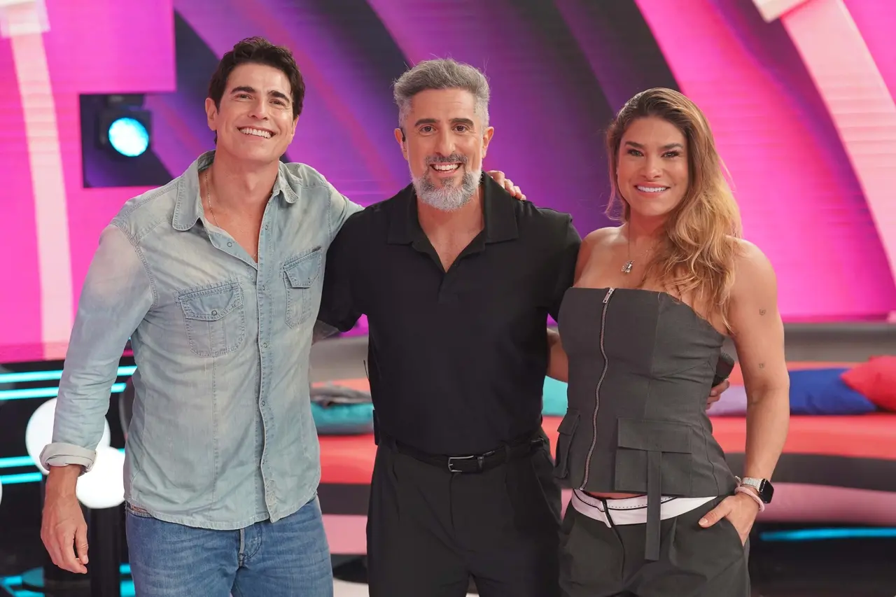 Reynaldo Gianecchini, Marcos Mion e Priscila Fantin sorriem abraçados no palco iluminado do programa Caldeirão com Mion. O trio compõe uma cena alegre em um cenário vibrante com tons de rosa e roxo ao fundo.