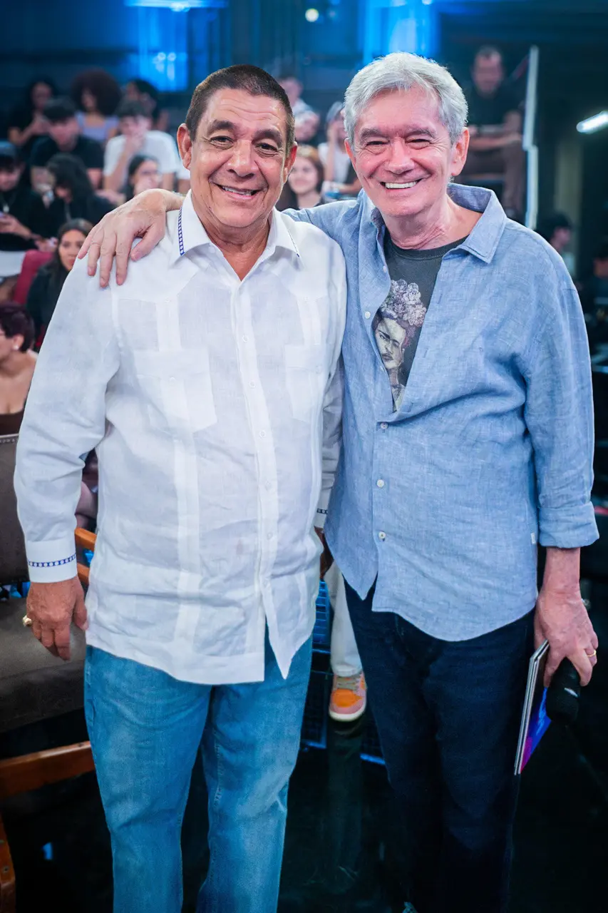 Zeca Pagodinho e Serginho Groisman sorriem abraçados no palco do programa 'Altas Horas', com a plateia visível ao fundo sob luzes azuis. Zeca veste uma camisa branca de botões e Serginho usa uma camisa azul clara sobre uma camiseta estampada, em um registro de amizade e celebração.