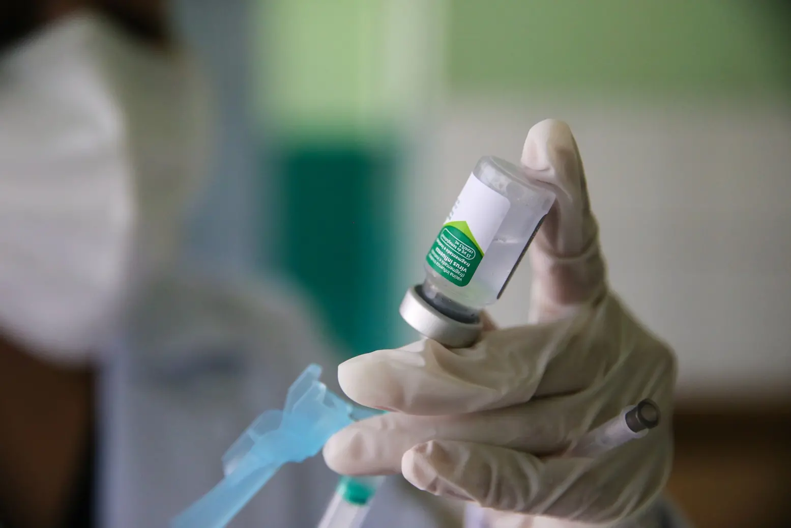 Mão enluvada de profissional de saúde preparando um frasco de vacina ou medicamento com uma seringa em um posto de saúde de Fortaleza.