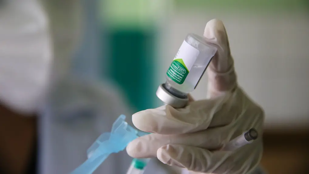 Mão enluvada de profissional de saúde preparando um frasco de vacina ou medicamento com uma seringa em um posto de saúde de Fortaleza.