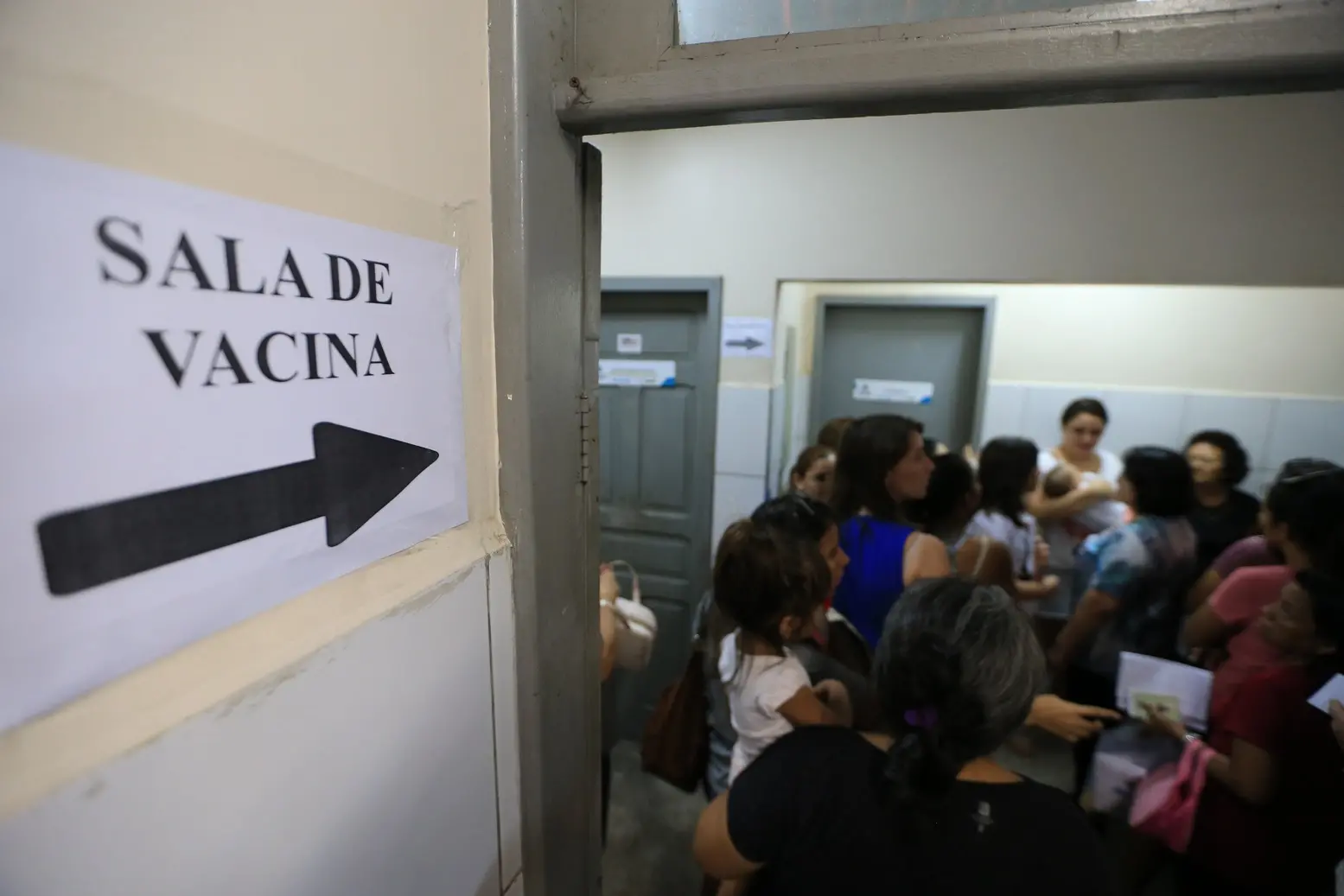 Multidão em um corredor de um posto de saúde, com um foco no sinal SALA DE VACINA indicando o caminho, sugerindo uma fila ou área de espera para imunização.