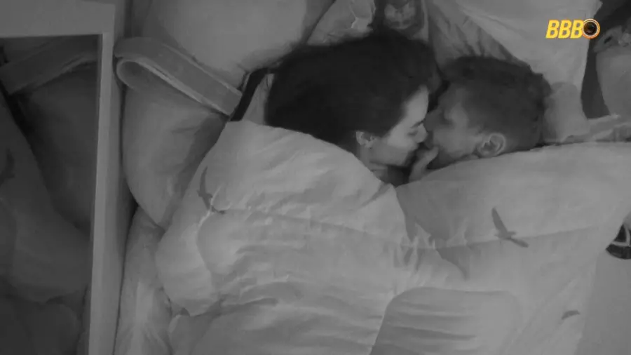 Imagem em preto e branco de um casal deitado na cama sob o edredom, trocando carinho e se beijando, com o logo do BBB no canto superior.