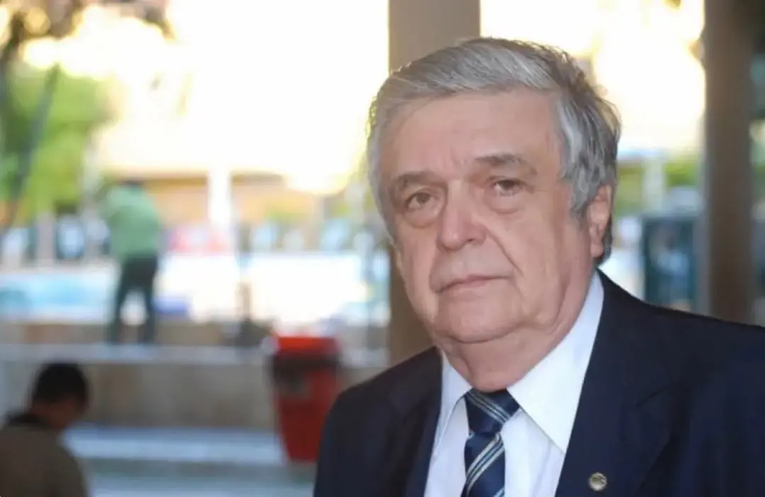 Homem idoso de cabelos grisalhos, usando terno e gravata, em ambiente externo desfocado, olhando em direção à câmera com expressão séria.