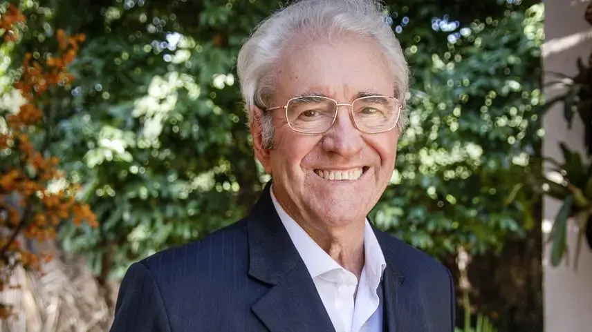 Juca de Oliveira. Homem idoso sorridente, de cabelo branco e usando óculos, vestido com terno escuro e camisa branca, em pé ao ar livre com árvores e folhagem verde ao fundo.