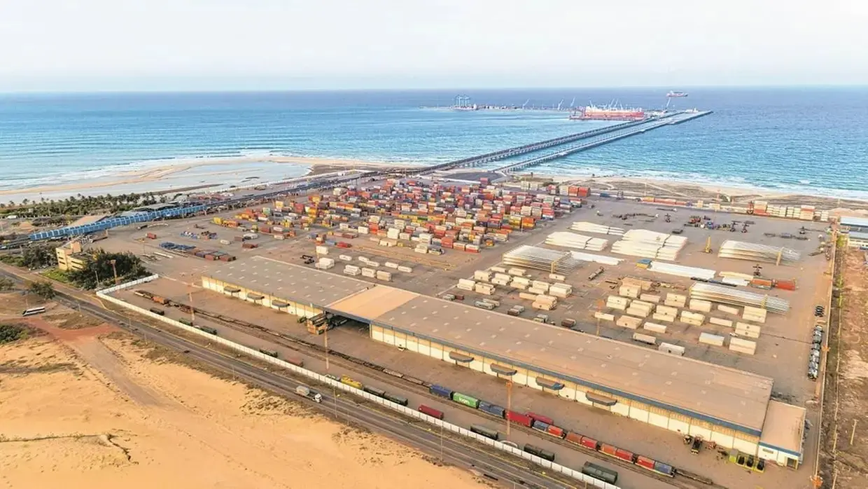Vista aérea do Porto do Pecém, um porto industrial à beira-mar, com grande área de contêineres coloridos organizados, armazéns, trilhos de trem e um longo píer que se estende até o oceano, onde há navios atracados.