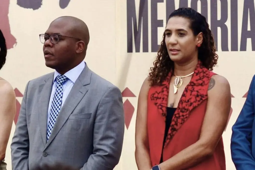 Duas pessoas em pé diante de um fundo com texto; um homem de terno cinza e gravata e uma mulher com cabelo cacheado usando colete vermelho, ambos olhando para o lado com expressão séria.