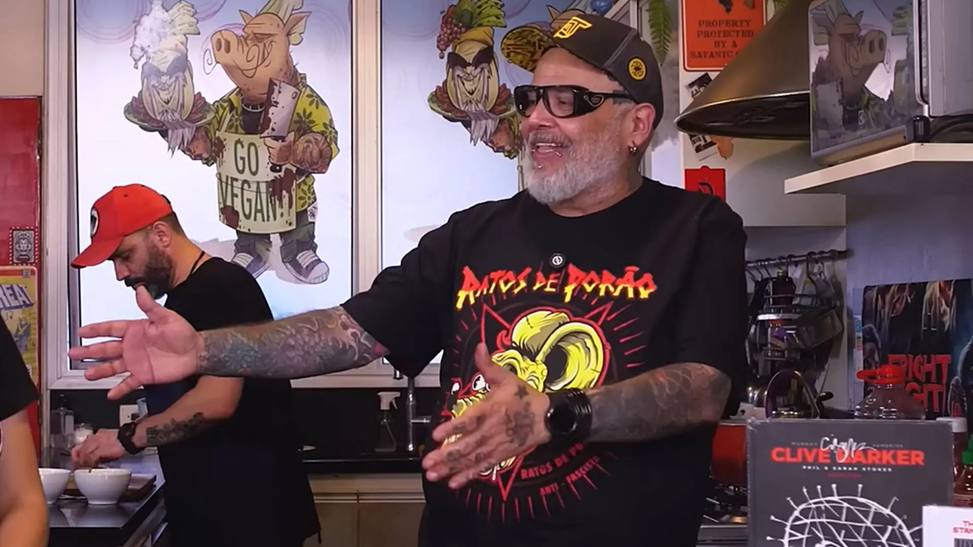 Homem de barba grisalha, usando óculos escuros, boné e camiseta preta da banda Ratos de Porão, gesticula com os braços abertos enquanto fala em uma cozinha. Ao fundo, há ilustrações de personagens com cabeças de animais e um homem de boné vermelho prepara algo sobre a bancada. À direita, aparecem prateleiras com objetos, um exaustor e um livro exposto.
