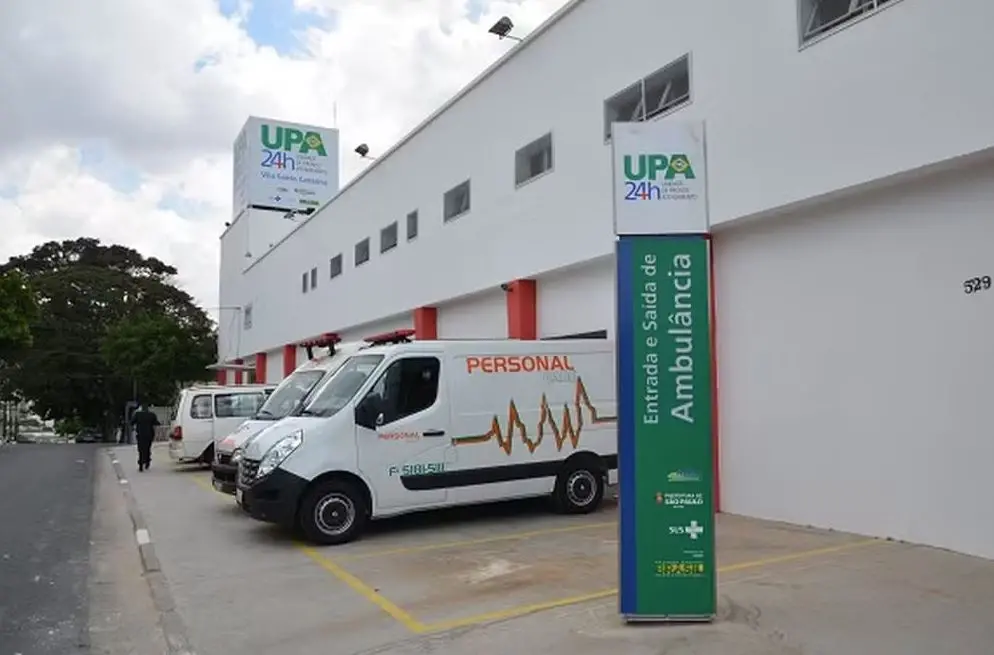 Fachada de uma Unidade de Pronto Atendimento (UPA 24h). Na área externa, há duas ambulâncias brancas estacionadas, uma delas com a palavra “PERSONAL” e um gráfico de batimentos cardíacos estampados na lateral. À direita, existe uma placa vertical indicando “Entrada e saída de ambulância”, com logotipos do SUS, Ministério da Saúde e Governo Federal. O prédio é branco, com janelas retangulares pequenas e um letreiro superior com o logo da UPA 24h e o texto Vila Santa Catarina.