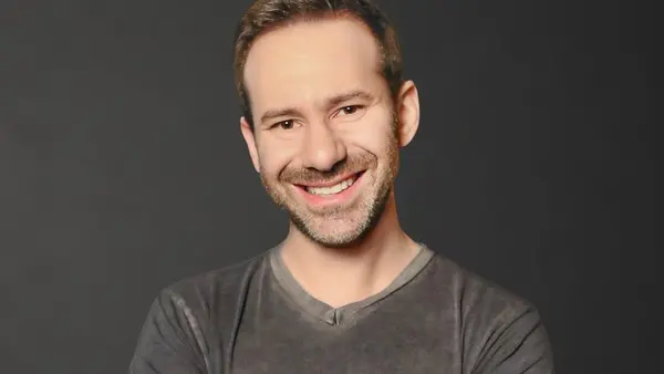 Imagem de um homem sorridente com barba e vestindo camiseta cinza, posando contra fundo escuro, transmitindo simpatia e confiança.