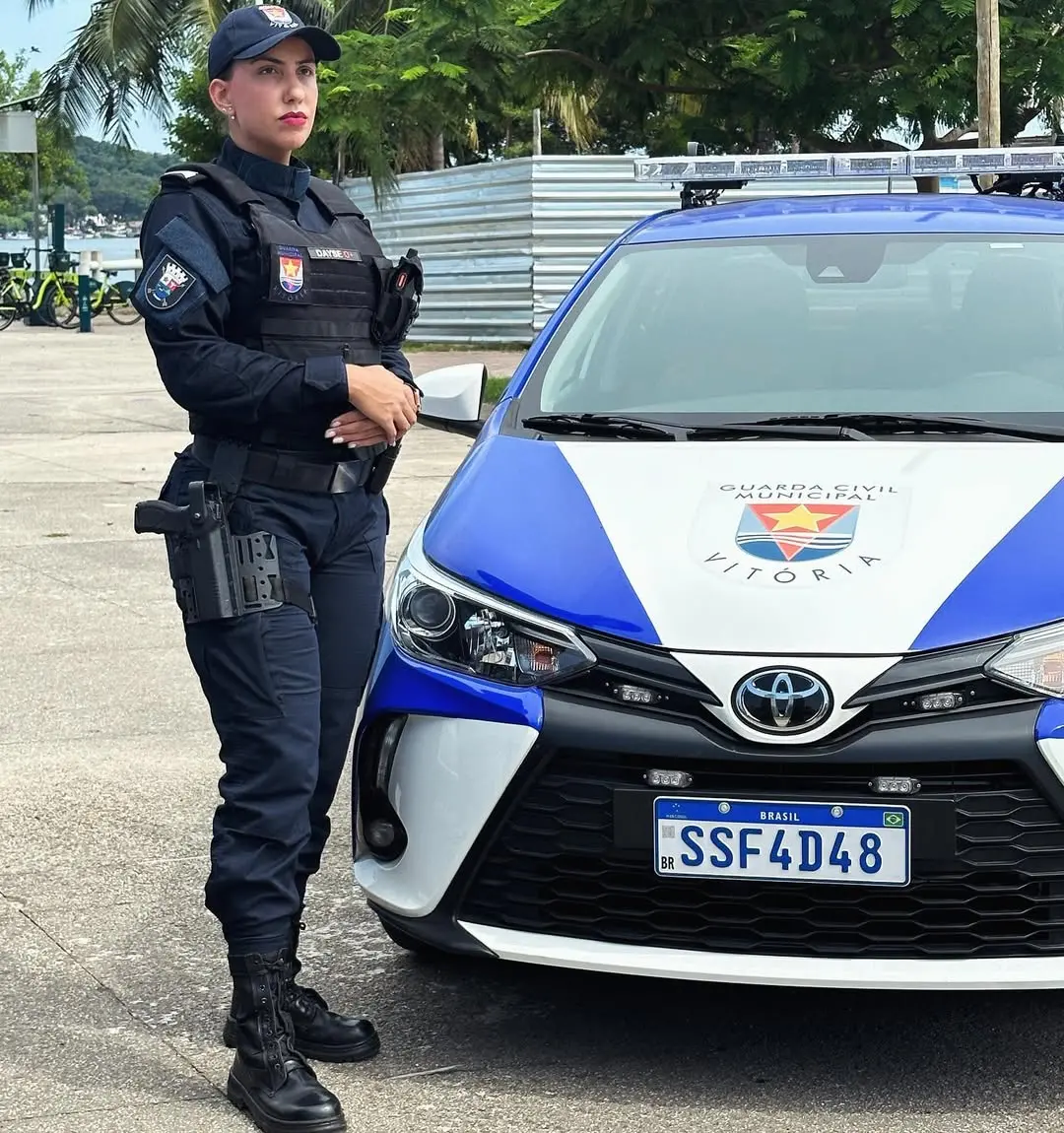 Policial feminina da Guarda Municipal de Vitória ao lado de viatura policial, vestida com uniforme azul escuro, capacete e equipamentos de segurança, em ambiente ao ar livre.