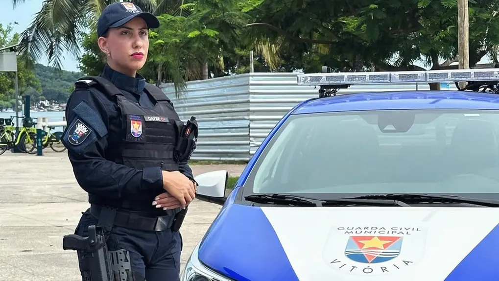 Policial feminina da Guarda Municipal de Vitória ao lado de viatura policial, vestida com uniforme azul escuro, capacete e equipamentos de segurança, em ambiente ao ar livre.