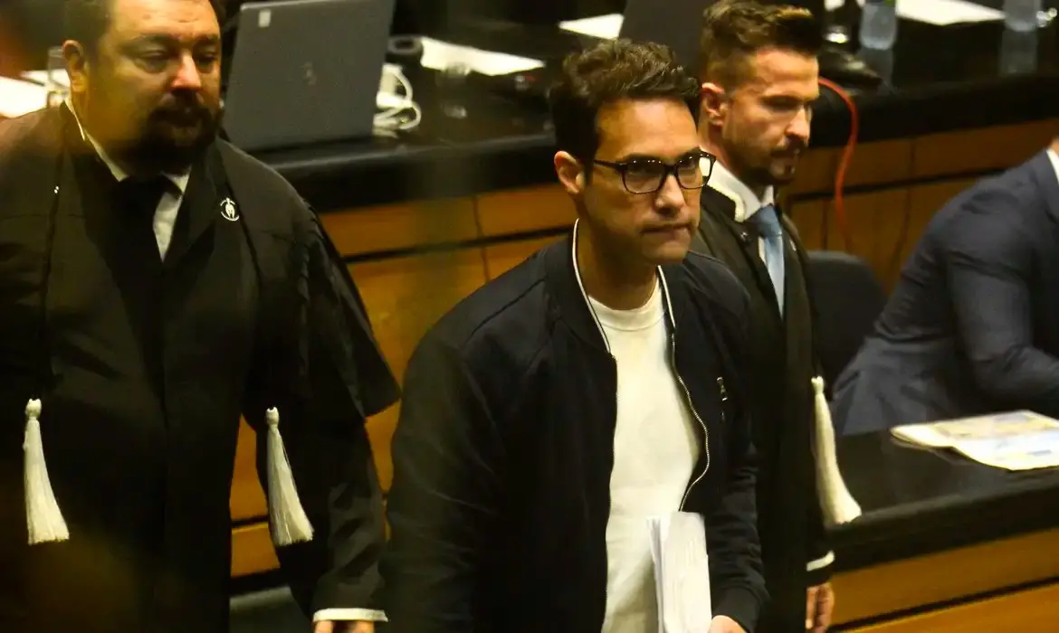 O réu Jairo Souza Santos Júnior, vestindo jaqueta preta e óculos, caminha em um tribunal ladeado por advogados em becas pretas durante o julgamento do caso Henry Borel. O ambiente ao fundo mostra a estrutura de madeira do plenário e equipamentos de escritório sob iluminação amarela.