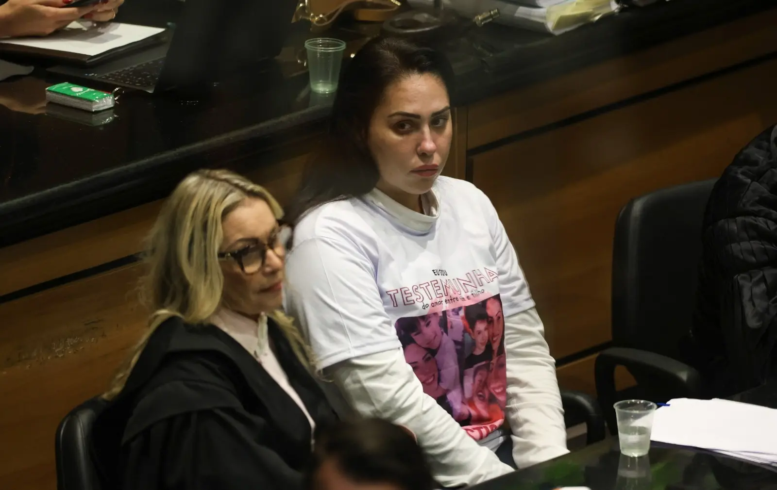 Monique é uma mulher branca de cabelo longo, liso e preto. Na foto, ela está ao lado da advogada de defesa.