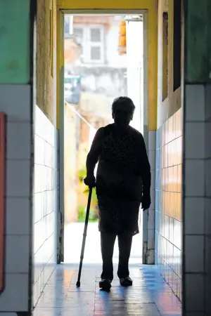 Sombra de idosa de costas, caminhando por um corredor, usando uma bengala. Ela vai em direção a rua em um dia ensolarado.