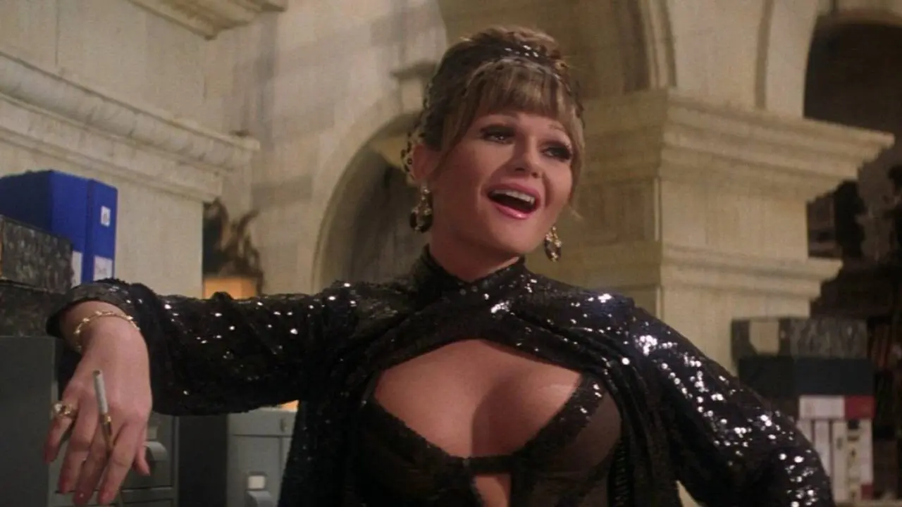 Mulher sorridente e elegante, com cabelo preso e franja, usando um vestido preto brilhante com decote e transparência. Ela está apoiada de forma descontraída em um móvel, segurando um cigarro na mão, em um ambiente interno que lembra uma casa ou escritório sofisticado, com paredes claras e detalhes clássicos ao fundo.