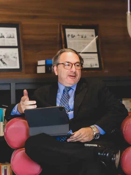 O advogado Roberto Podval gesticula enquanto segura um tablet, sentado em uma poltrona vermelha em um escritório com fundo de madeira e quadros decorativos. Ele veste um terno escuro com camisa azul e gravata estampada, compondo uma cena de trabalho intelectual e conversa dinâmica.