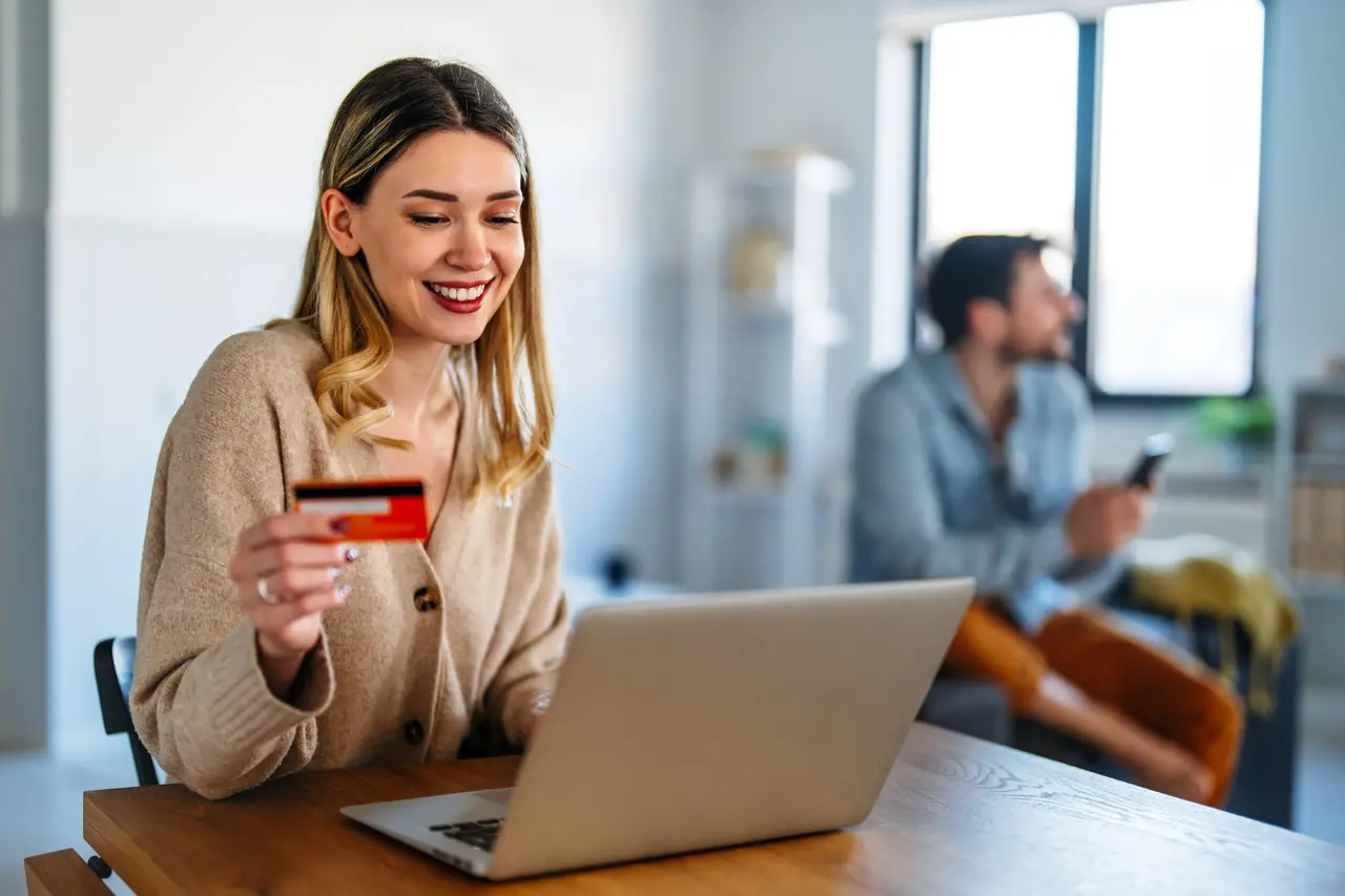 Mulher sorridente segura um cartão de crédito enquanto usa um notebook em uma mesa; ao fundo, um homem desfocado mexe no celular em um ambiente doméstico iluminado por luz natural.