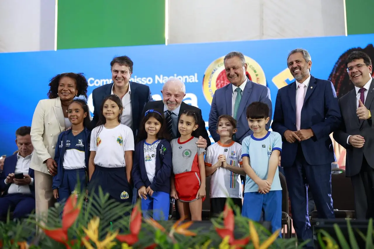 Grupo de autoridades e crianças posam para foto em um palco durante evento oficial. Ao centro, um homem idoso de barba branca, sorridente, inclina-se levemente entre as crianças. Ao lado dele, outros homens e uma mulher, todos bem vestidos com trajes formais, também sorriem. Na frente, cerca de oito crianças usam uniformes escolares coloridos e variados, algumas com brasões ou logotipos. Ao fundo, há um painel azul com elementos gráficos e texto parcialmente visível, além de bandeiras e símbolos institucionais. Na parte inferior da imagem, aparecem plantas ornamentais com flores tropicais.