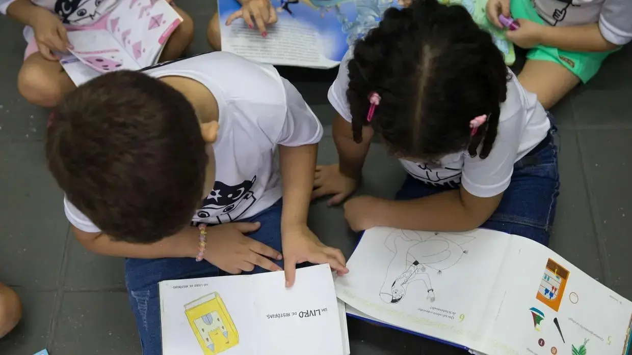 Crianças sentadas no chão de uma sala de aula, vestindo camisetas brancas, olham atentamente para livros coloridos abertos em suas mãos.