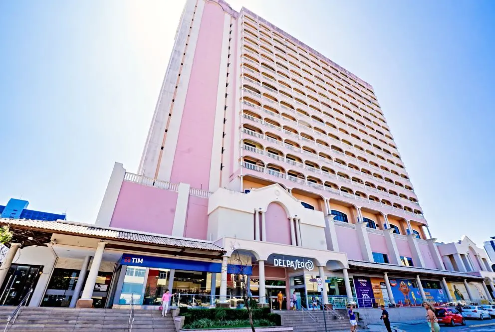 A fachada do Shopping Del Paseo em Fortaleza apresenta sua característica torre comercial de tons rosados e múltiplas varandas sob um céu azul claro. No nível da rua, destacam-se a entrada principal com o letreiro do shopping, lojas como a Tim e a movimentação de pedestres na calçada e escadarias.