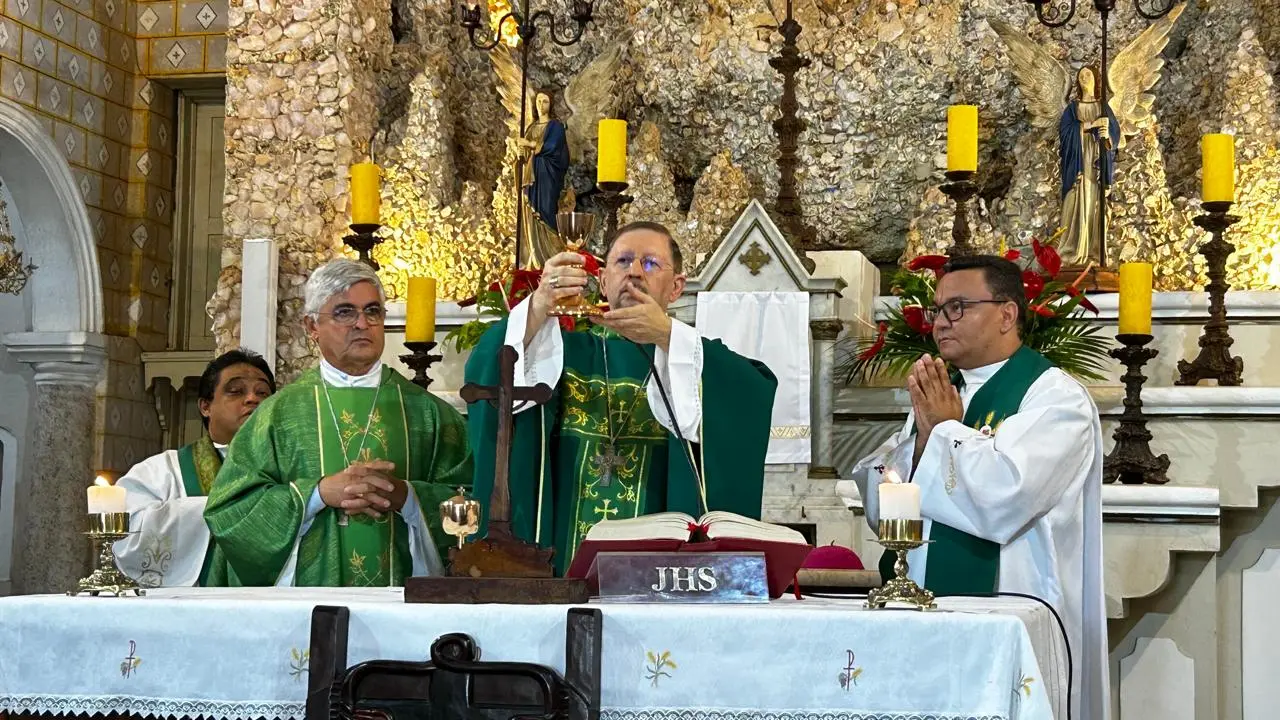 Dom Luís Pepeu celebra uma missa em um altar de pedras rústicas, elevando o cálice sagrado. Ele veste paramentos verdes com detalhes dourados e está acompanhado por outros religiosos. O ambiente é decorado com velas amarelas acesas e estátuas de anjos ao fundo.