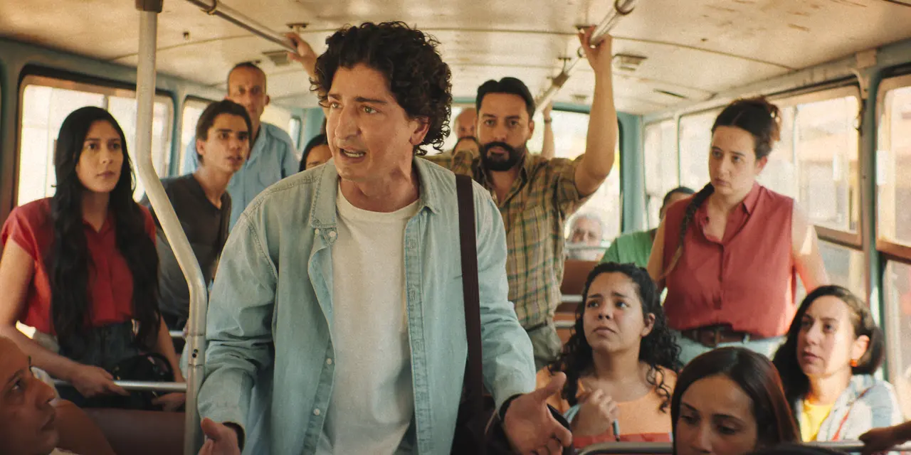 Interior de um ônibus lotado, com várias pessoas sentadas e em pé. No centro da imagem, uma pessoa de cabelos cacheados e camisa clara está de pé no corredor, gesticulando com as mãos enquanto fala. As outras pessoas ao redor olham na direção desta pessoa. A iluminação é natural, vinda das janelas laterais, e o ambiente aparenta ser de um ônibus antigo, com barras metálicas e bancos simples.