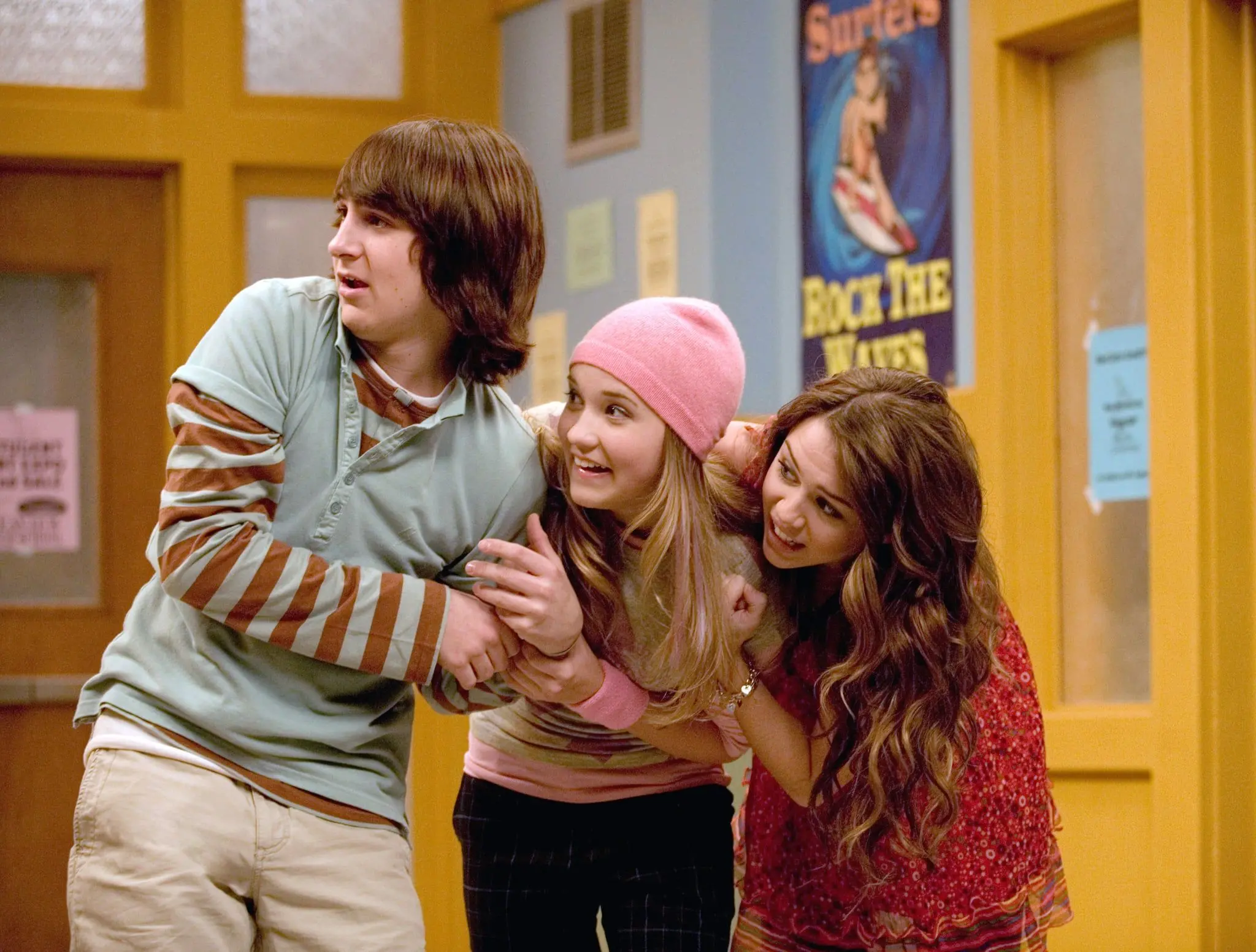 Oliver (Mitchel Musso), Lilly (Emily Osment) e Miley (Miley Cyrus) em imagem de divulgação da série 'Hannah Montana'. Na iagem, o trio aparece em um corredor de escola, enquanto olham para o lado com expressões de surpresa e alegria. O cenário apresenta tons de bege e amarelo com cartazes ao fundo, compondo uma cena divertida e dinâmica da série.