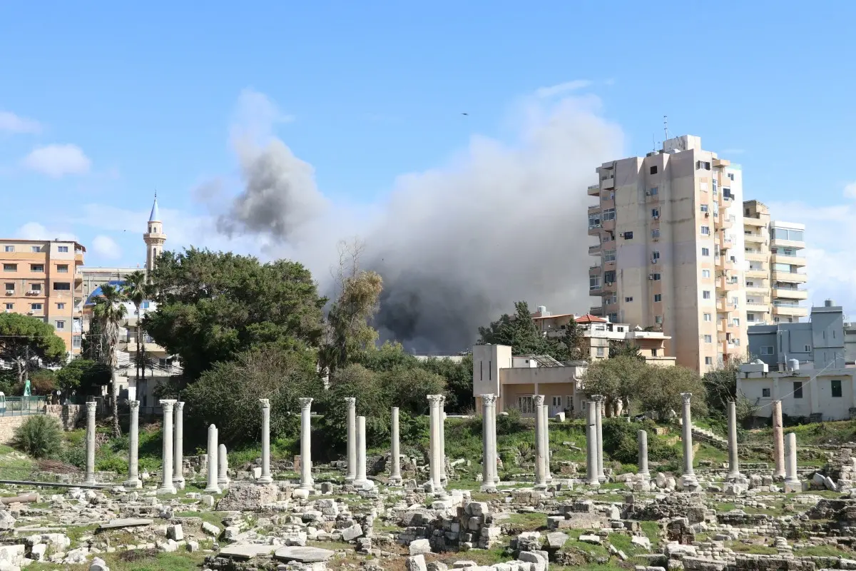 Incêndio e fumaça escura se elevando ao céu em uma área urbana com construções modernas e ruínas ao fundo.