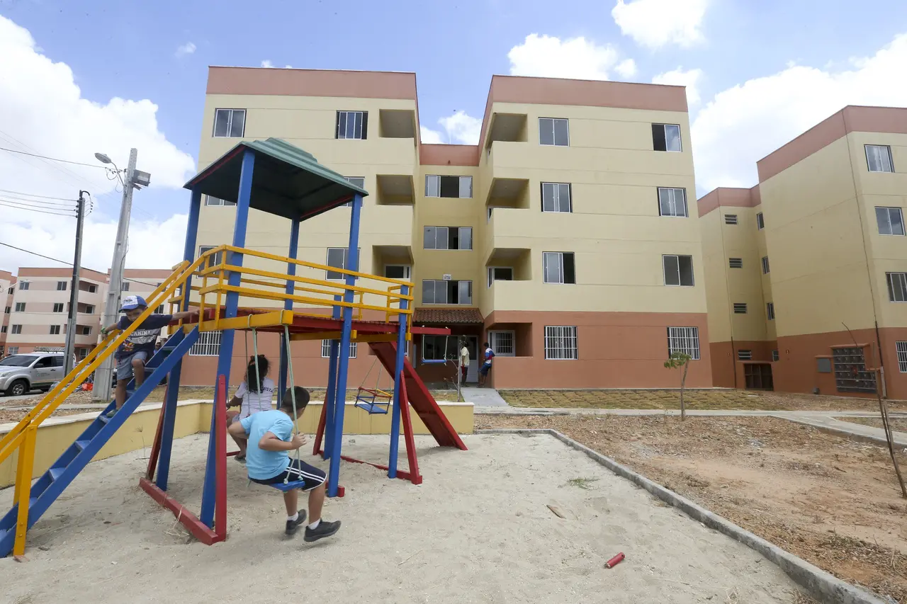 Crianças brincam em um parquinho colorido de areia em frente aos blocos de apartamentos do Residencial Luiz Gonzaga, que apresentam fachadas em tons de bege e terracota. A cena captura um momento cotidiano de lazer sob um céu ensolarado, destacando a infraestrutura da área habitacional de Fortaleza.