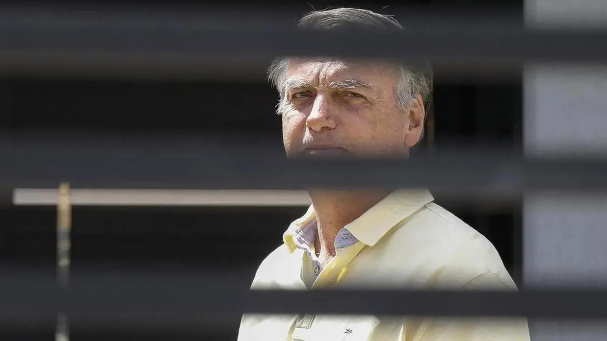 Bolsonaro aparece com expressão séria atrás de grades de sua residência.