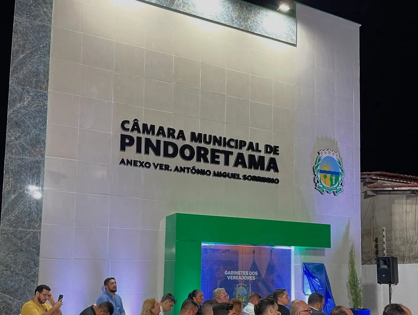 Fachada moderna da Câmara Municipal de Pindoretama, com revestimento claro e detalhes em pedra. No centro, o nome “Câmara Municipal de Pindoretama” aparece em letras escuras, acompanhado da identificação do anexo. À direita, há o brasão do município iluminado. Na parte inferior, um grupo de pessoas está sentado em cadeiras, assistindo a um evento, enquanto um pequeno espaço com iluminação verde destaca a entrada dos gabinetes dos vereadores. Ao lado, há uma caixa de som e estruturas de apoio, indicando uma cerimônia ou sessão pública.