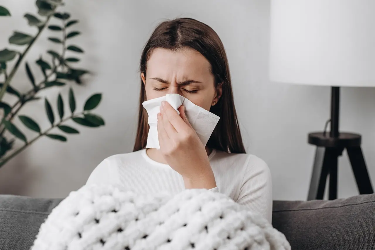 Mulher tossindo com sensação de resfriado ou gripe, cobrindo o nariz com lenço de papel, em ambiente de casa com plantas e luminária ao fundo.
