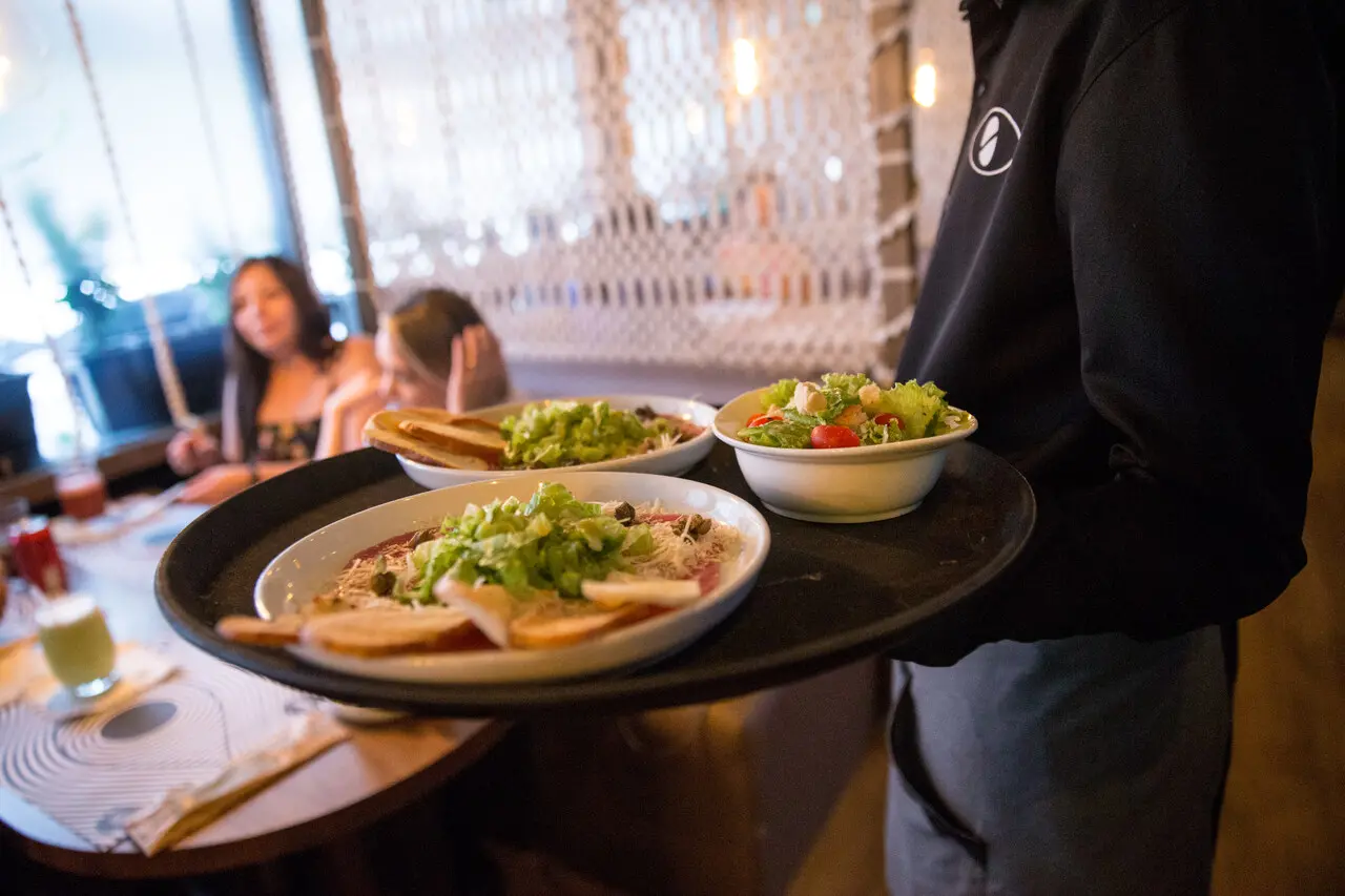 Garçom segurando bandeja com pratos de salada e entrada em restaurante, enquanto clientes aparecem ao fundo desfocados.