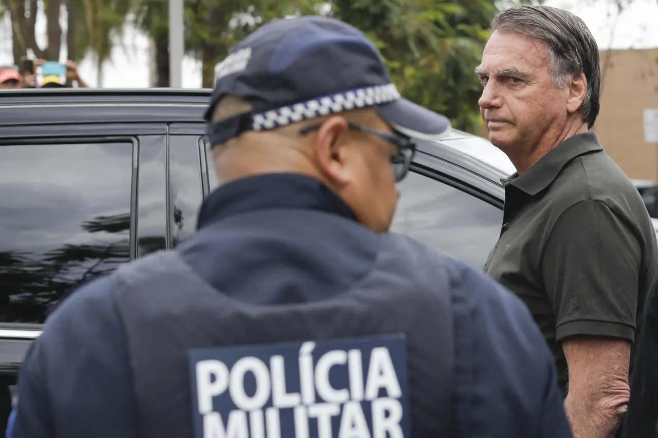Jair Bolsonaro observa seriamente em direção ao lado esquerdo da imagem, posicionado atrás de um policial militar que aparece de costas em primeiro plano. A composição destaca o contraste entre a autoridade policial e a expressão atenta do ex-presidente ao lado de um veículo preto.
