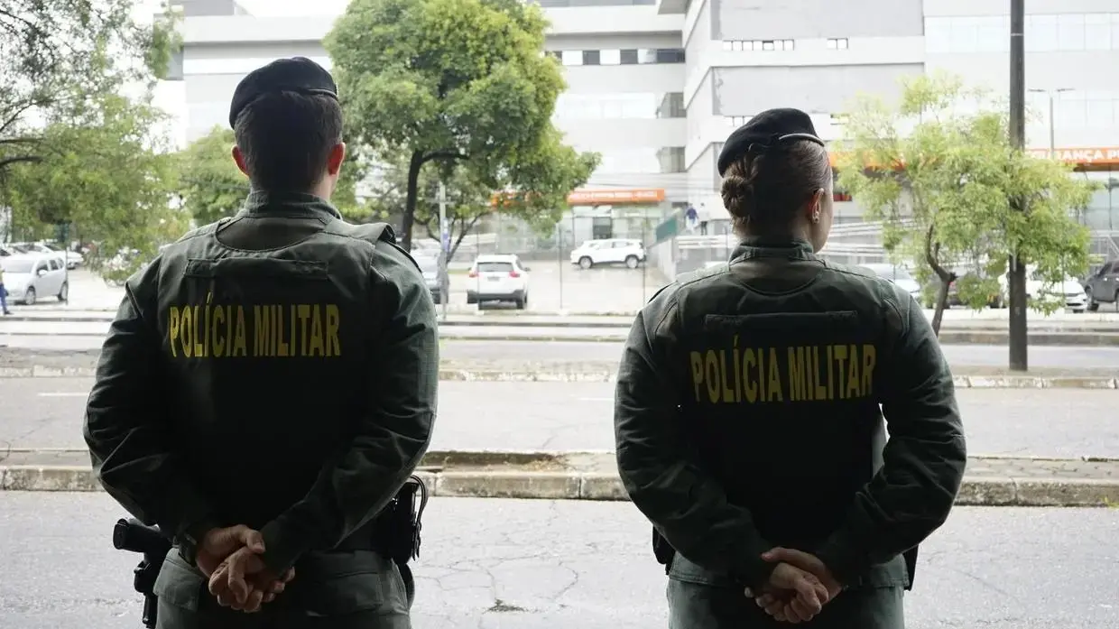 Dois policiais militares, um homem e uma mulher, estão de costas, usando fardas verdes com a inscrição “Polícia Militar” em amarelo. Ambos mantêm as mãos para trás, em posição de descanso. Eles estão em uma calçada, diante de uma rua com carros passando e árvores, com prédios ao fundo em um ambiente urbano.