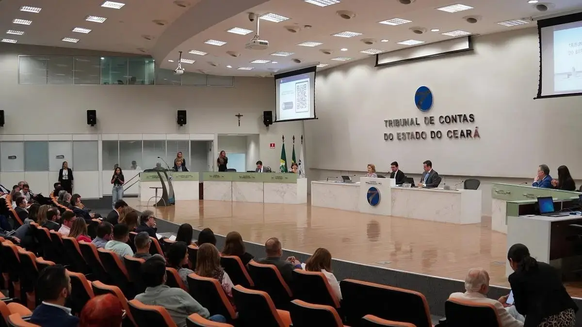 Registro de sessão ou evento no plenário do TCE-CE. A imagem mostra o público em poltronas laranjas e pretas em primeiro plano, voltados para a mesa de autoridades ao fundo. Na parede branca central, destaca-se o letreiro azul e vermelho: 