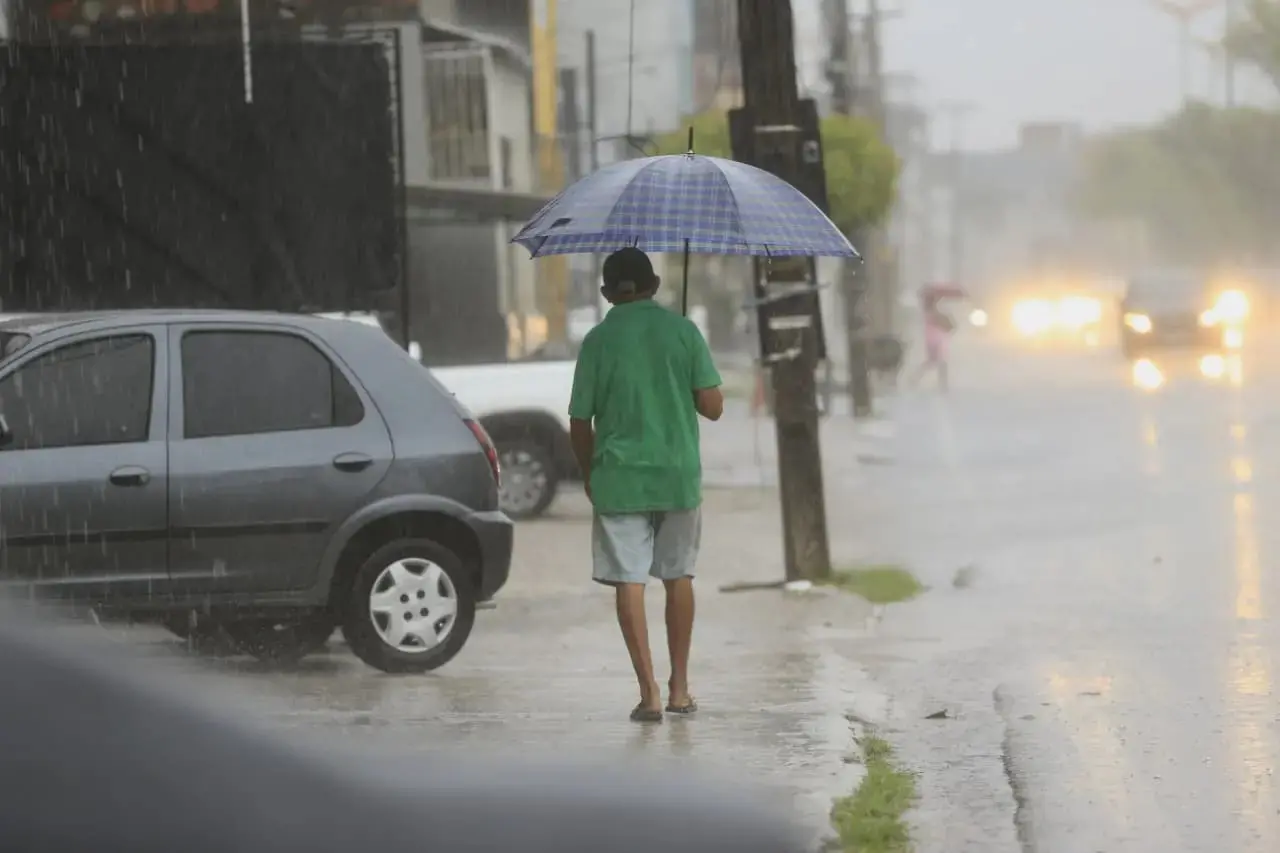 foto de um senhor de costas, com blusa verde e bermuda branca, caminhando com guarda chuva aberto em calçada em um dia de chuva intensa.