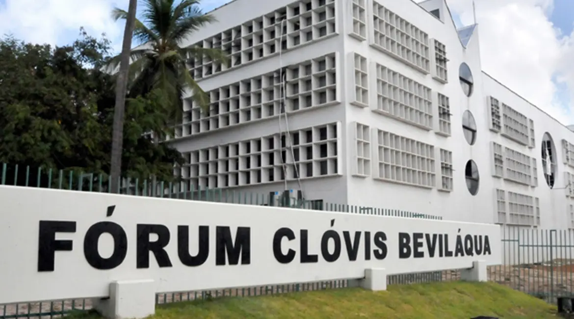 Fachada branca do Fórum Clóvis Beviláqua, com arquitetura modernista repleta de janelas retangulares e circulares. Em primeiro plano, uma placa de identificação branca com o nome do prédio em letras pretas.