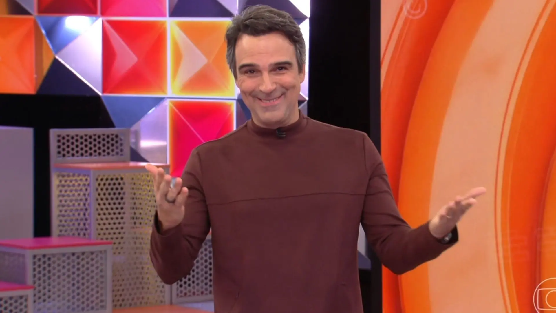 Apresentador de televisão sorridente durante programa de TV com fundo colorido em tons de laranja, roxo e geométrico, em estúdio de gravações.