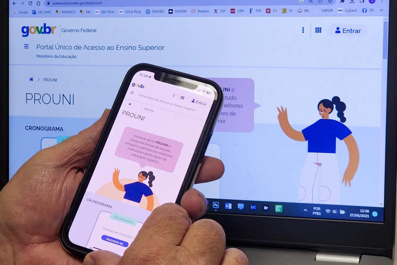 Pessoa segura um smartphone exibindo a página do Prouni, com o portal gov.br aberto ao fundo em um computador portátil.