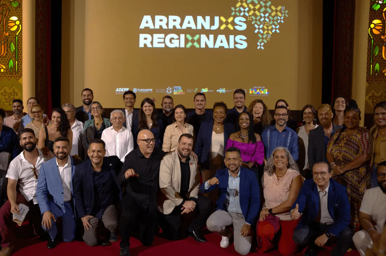 Um grupo diverso de pessoas sorri e posa para uma foto coletiva em um palco, com alguns integrantes agachados à frente e outros em pé ao fundo sobre um tapete vermelho. Atrás deles, um telão exibe a logomarca 'Arranjos Regionais' junto a um mapa do Brasil estilizado e marcas institucionais do Governo Federal.
