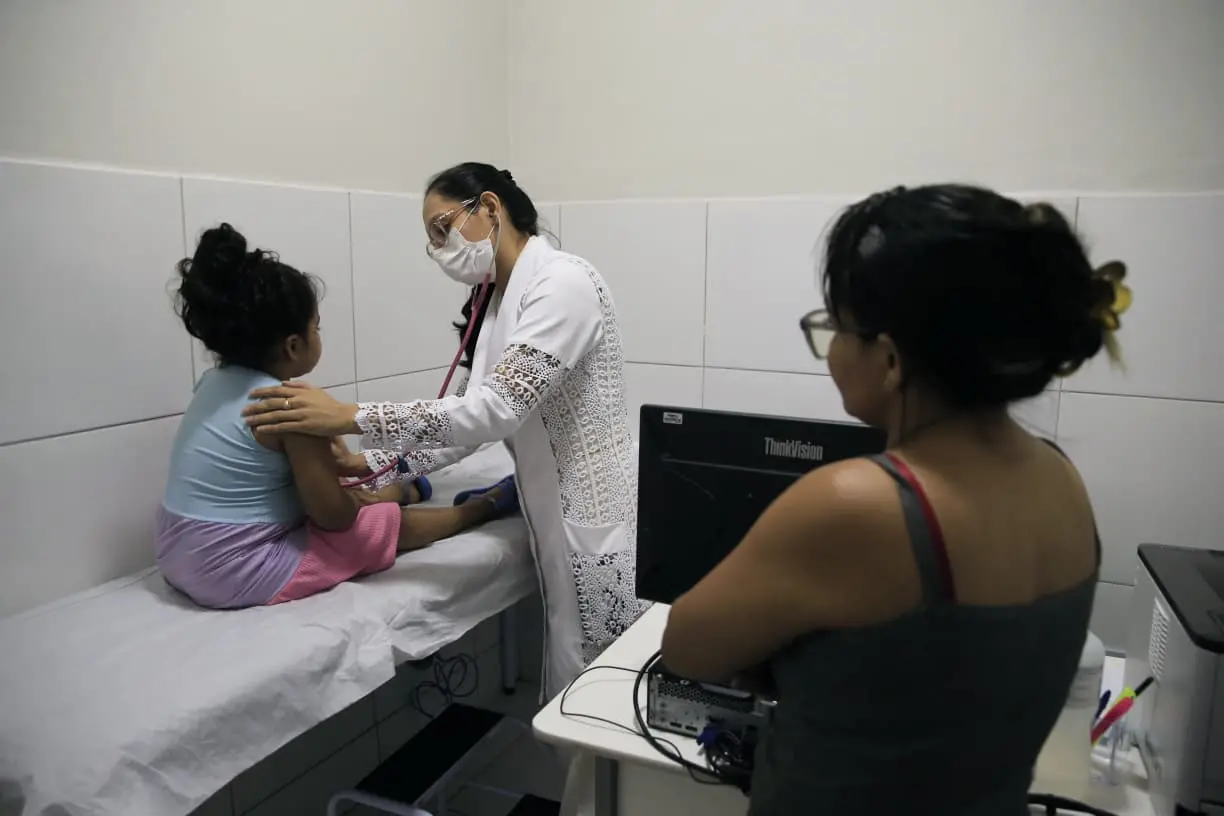 criança de costas, sendo examinada por profissional de saúde. ao lado, uma mulher de costas observa. o espaço é uma sala de atendimento de saúde.