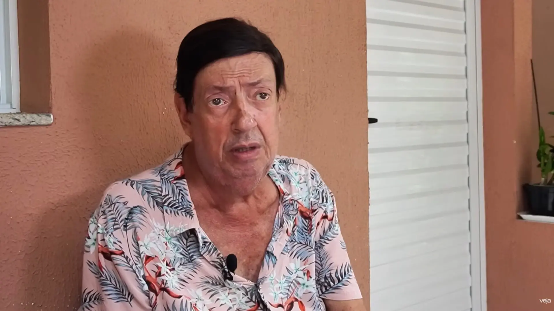 O ator Marcos Oliveira aparece em um plano médio, vestindo uma camisa de botão rosa com estampa tropical de folhagens e flores. Ele está sentado em uma área externa com paredes de tom terracota e uma porta de metal branca ao fundo, mantendo uma expressão séria enquanto fala.