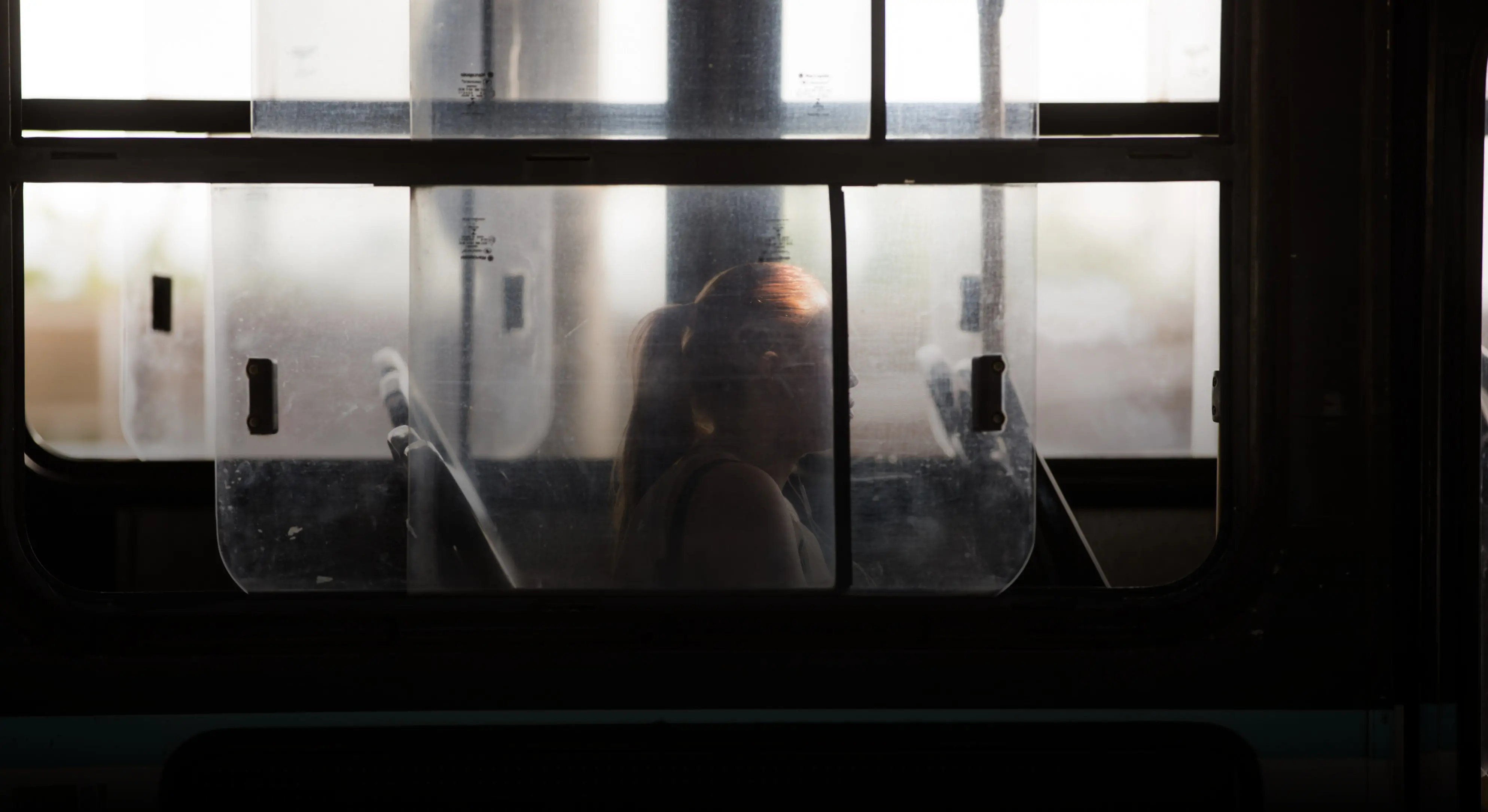 Silhueta de mulher jovem com cabelo preso em um rabo de cavalo, vista através da janela de um ônibus urbano. A luz de fundo destaca seu contorno contra o vidro translúcido e a moldura escura.