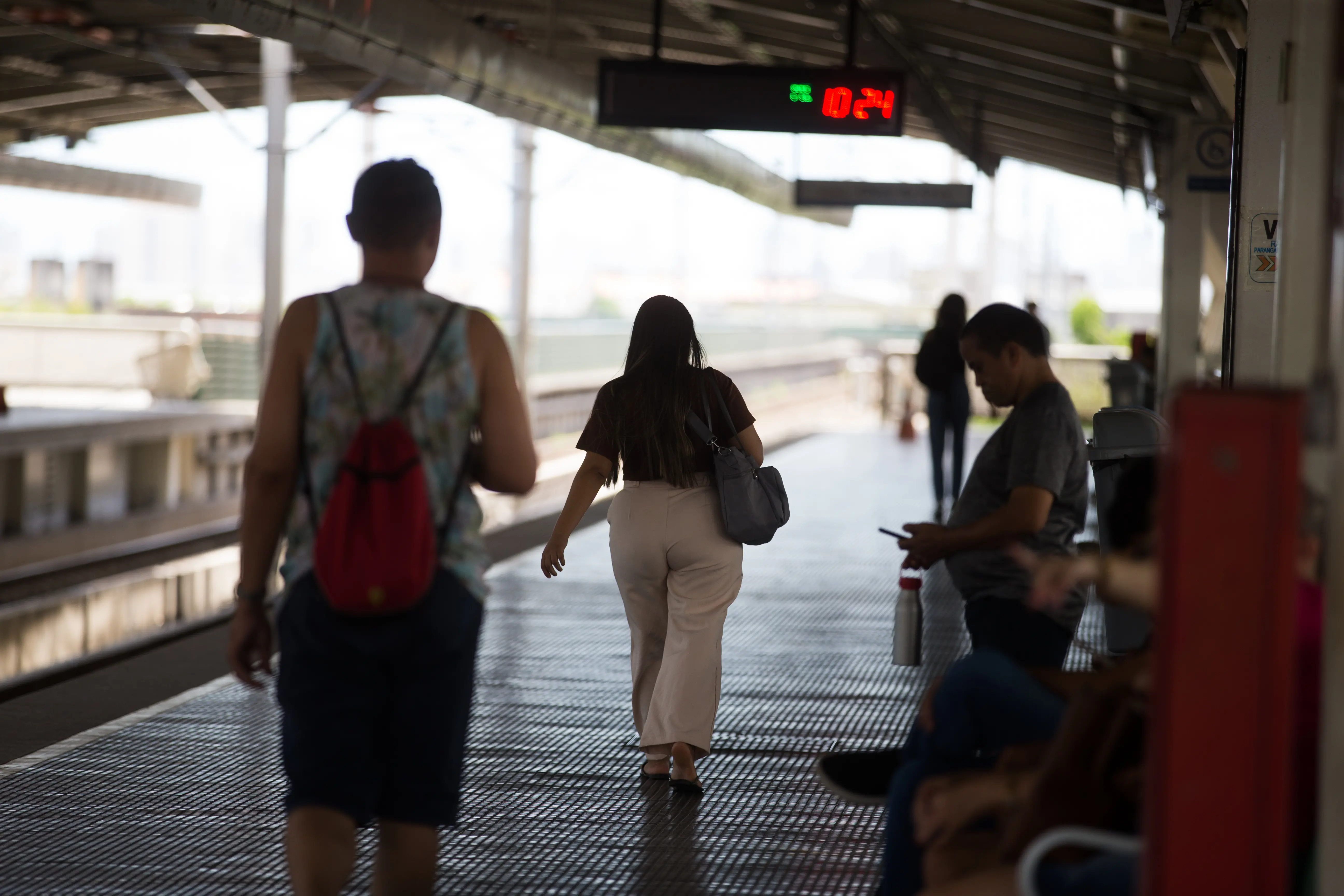 Mulher de costas caminha por plataforma de metrô em direção ao trem. Outros passageiros aguardam sob o abrigo da estação, com relógio digital ao fundo marcando 10:24.