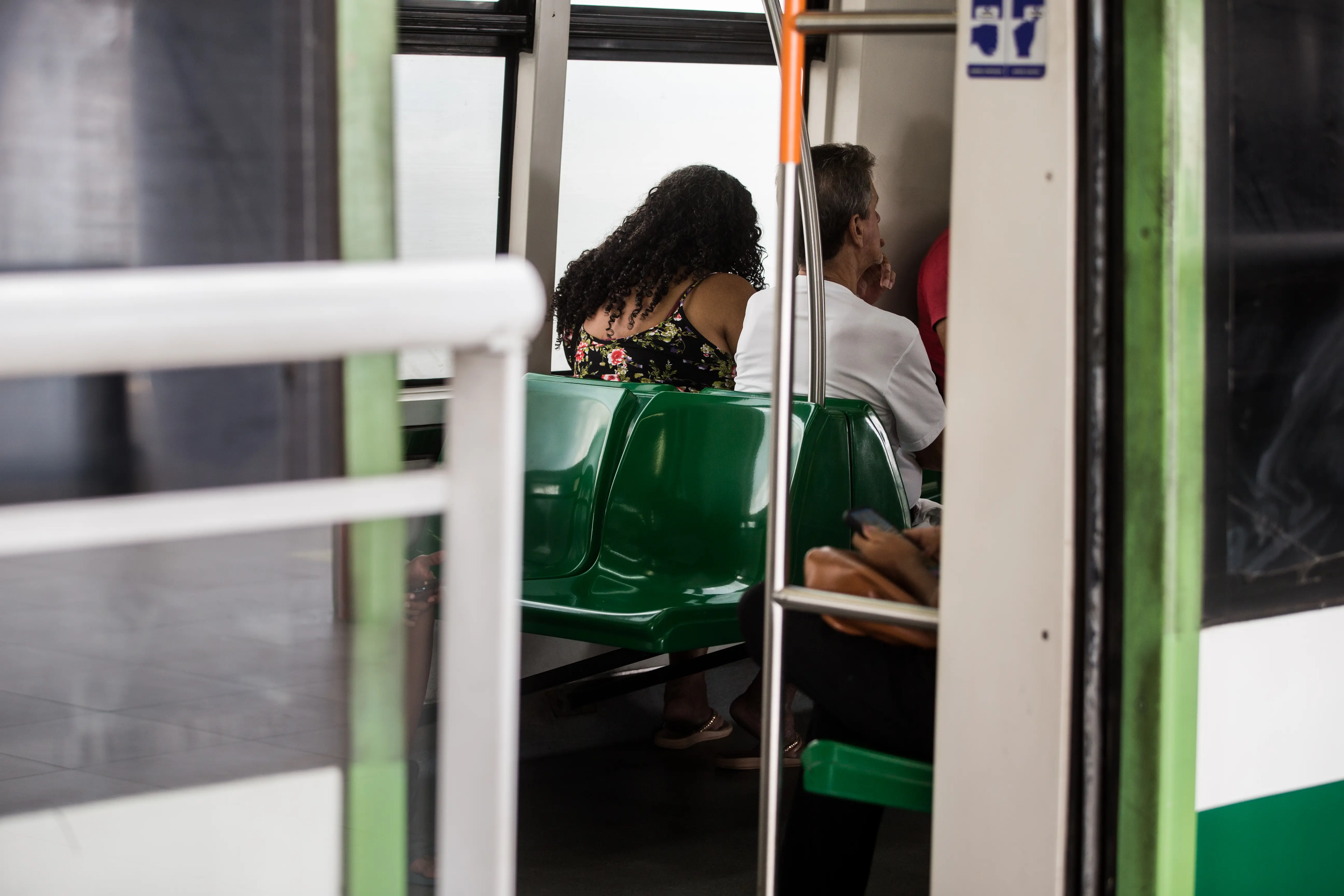Interior de vagão de metrô com assentos verdes. Uma mulher de cabelos cacheados e um homem estão sentados de costas, olhando pela janela.