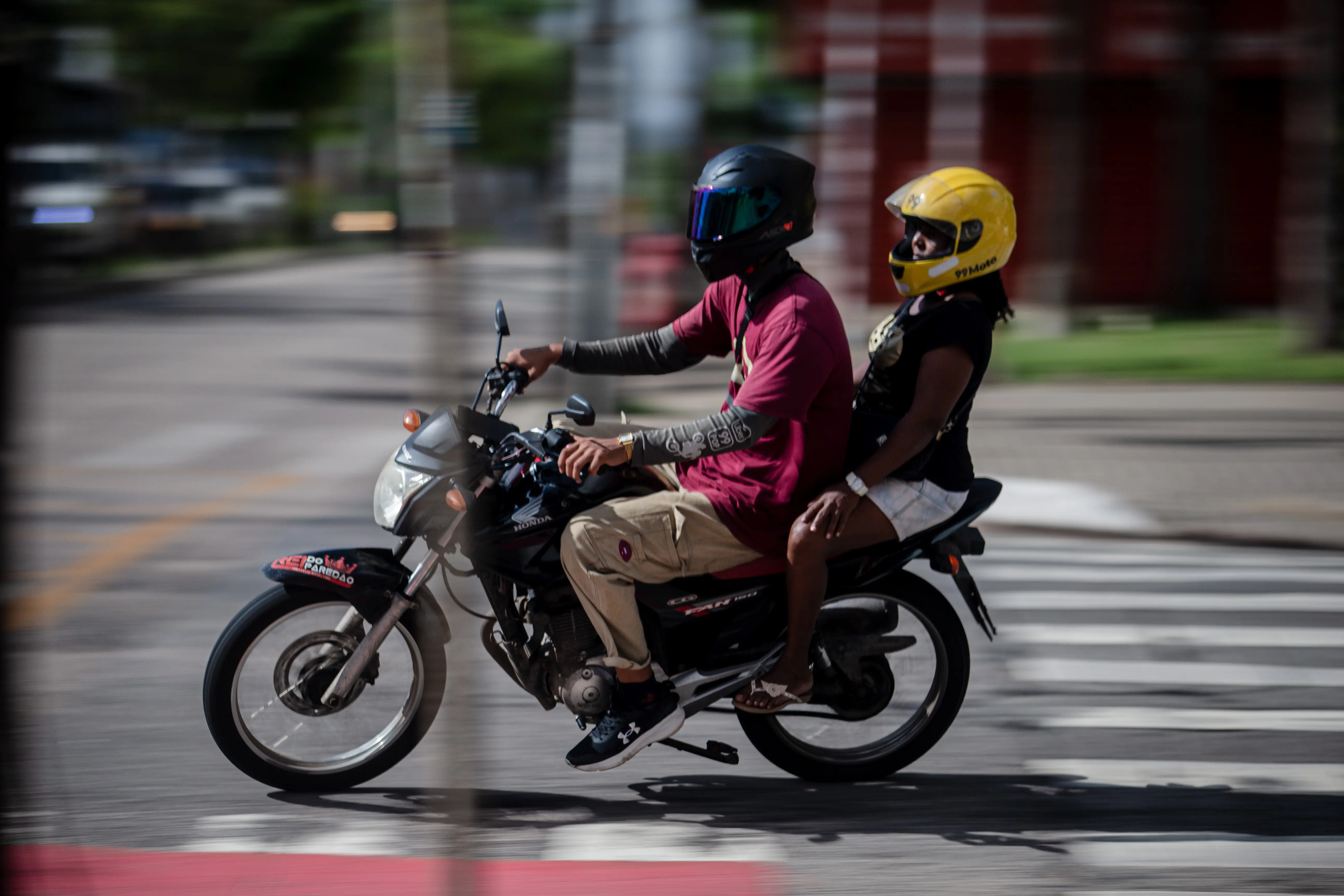 Moto preta atravessa faixa de pedestres com dois ocupantes. A passageira utiliza um capacete amarelo com o logotipo 99 Moto.