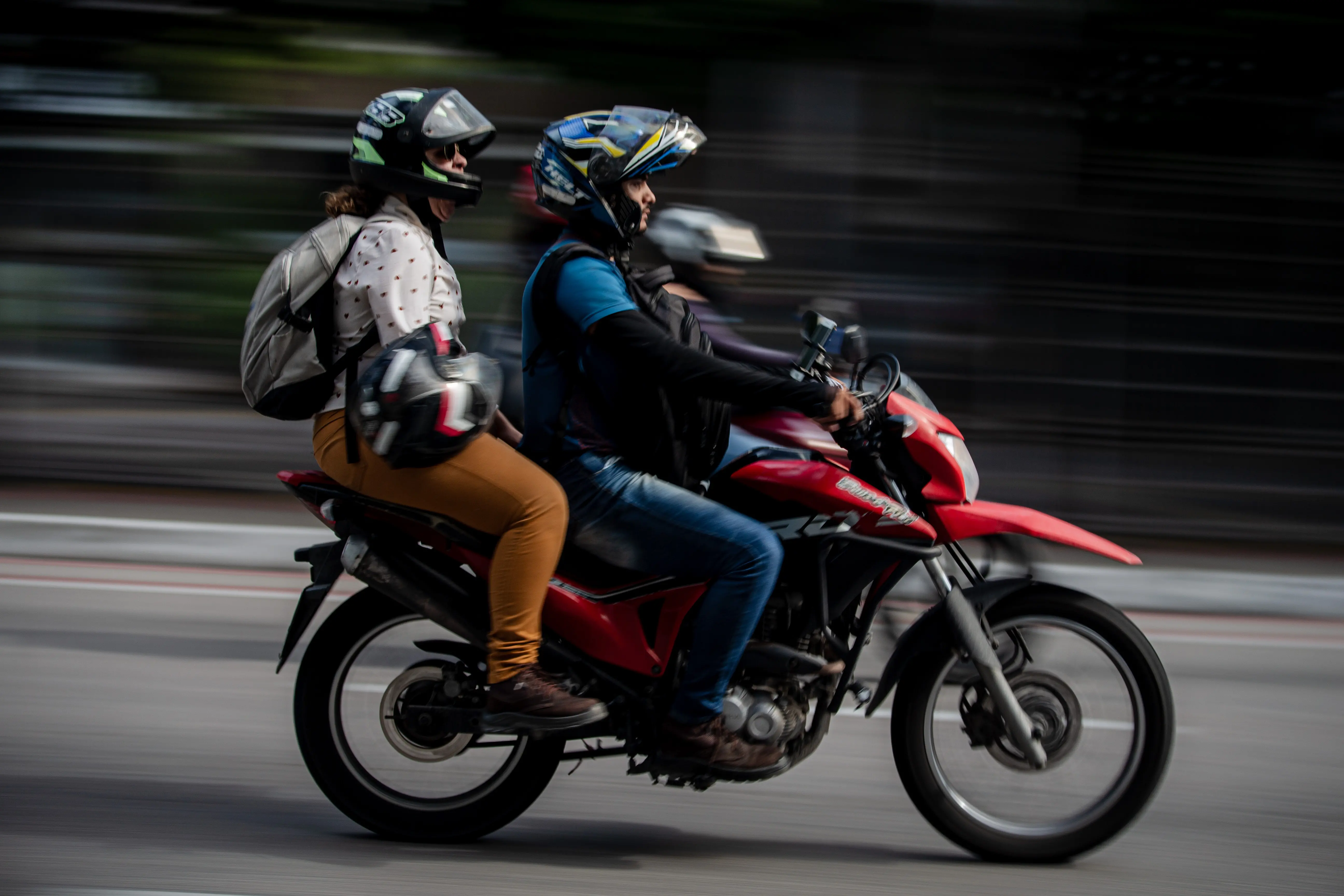 Motocicleta vermelha em movimento com piloto e passageira utilizando capacetes e mochilas. Efeito de velocidade ao fundo.