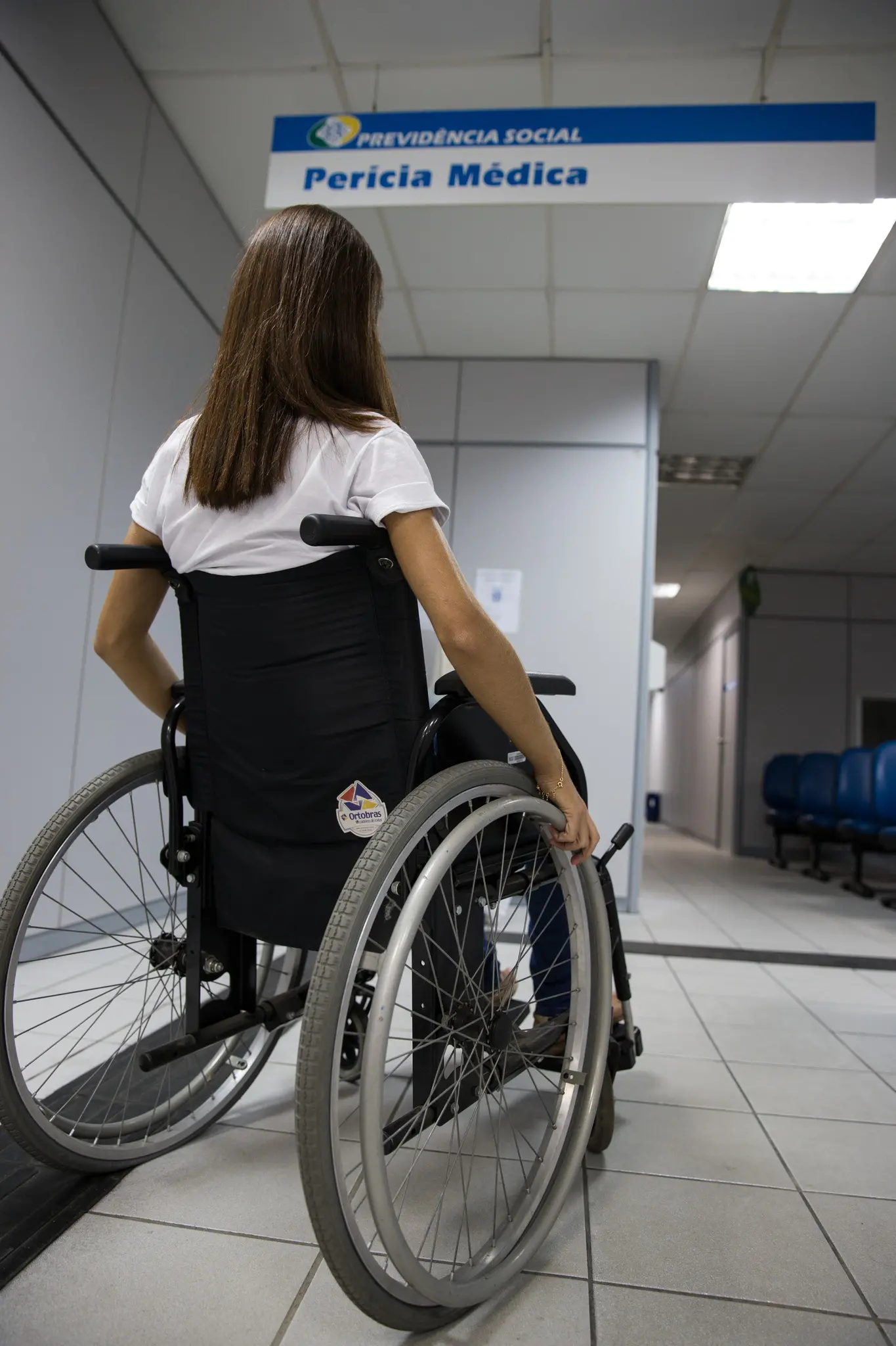 Mulher cadeirante em corredor, perto de placa que indica perícia médica.